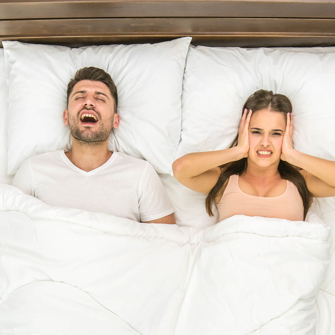 Man snoring in bed, wife covering her ears.